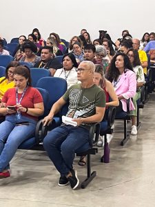 Para Todos Verem: várias pessoas sentadas em cadeiras azuis olhando para frente. 